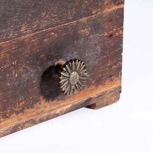 May include: A rectangular wooden box with a dark, weathered brown finish and visible wood grain. The front panel features a single drawer pull shaped like a metallic, antique brass-toned flower with textured, radiating petals.