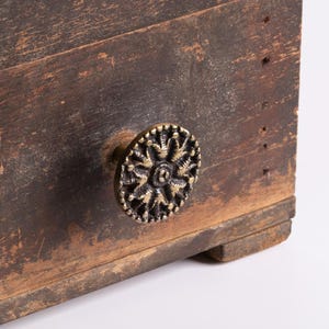 May include: A dark brown, weathered wooden drawer front featuring a circular, antique brass-toned metal knob. The knob has a textured, radial pattern with small, raised decorative details around its edge, attached to the centre of the wood.