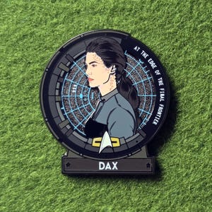May include: A circular enamel pin featuring a profile illustration of a character with dark hair and spotted markings on the neck and face. The background is a blue radar grid with the text AT THE EDGE OF THE FINAL FRONTIER and DAX at the base.