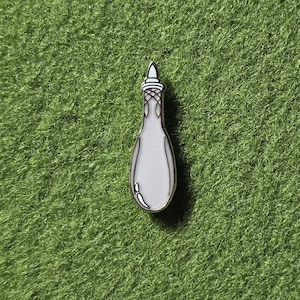 May include: A silver-colored enamel pin in the shape of a perfume bottle. The pin has a white center and a detailed top with a silver outline. The pin is set against a green background.