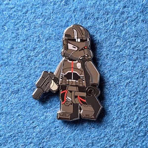 May include: A pin featuring a stylized, gray and black character in a helmet and armor, holding a blaster. The character has a skull emblem and red accents. The pin is set against a blue background.