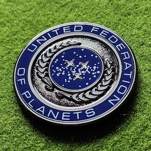 May include: A round, metallic coin with a blue and silver design. The coin features the words "UNITED FEDERATION OF PLANETS" around the edge. The center has a blue field with stars and a symbol.
