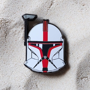 May include: A white and red enamel pin shaped like a clone trooper helmet. The helmet features a black visor, a vertical red stripe down the centre, and a black antenna attached to the left side. The design includes black outlines and details.