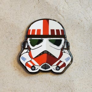 May include: A metal enamel pin shaped like a trooper helmet featuring a white base with red and dark red vertical stripes. The helmet has dark green visor lenses, black outlines, and detailed ventilation ports on the sides and front mouth area.