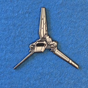 May include: A metal enamel pin shaped like a white and grey spacecraft with a central vertical wing and two angled side wings. The design features a dark cockpit window and detailed panel lines, capturing the appearance of a starship shuttle.
