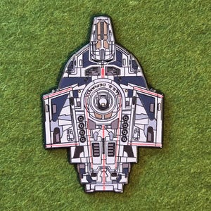 Puede incluir: Un pin de esmalte metálico con la forma de la nave espacial U.S.S. Defiant. El diseño presenta tonos gris claro y azul oscuro con detalles de paneles intrincados, un emblema circular central y finas líneas rojas a lo largo de la estructura.