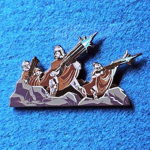 Pode incluir: Um alfinete de esmalte detalhado com um grupo de figuras em capas marrons e capacetes brancos, segurando armas grandes. O alfinete &eacute; colocado em um fundo de rochas cinzentas e um fundo azul.