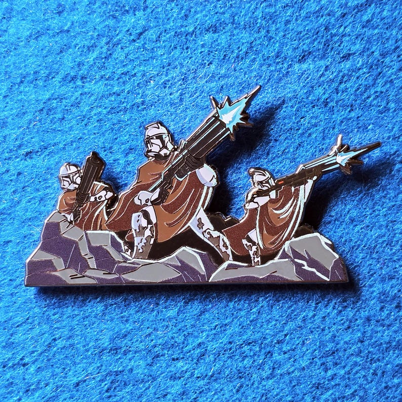 Pode incluir: Um alfinete de esmalte detalhado com um grupo de figuras em capas marrons e capacetes brancos, segurando armas grandes. O alfinete &eacute; colocado em um fundo de rochas cinzentas e um fundo azul.