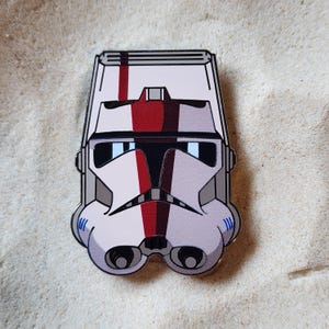 May include: A metal enamel pin shaped like a white clone trooper helmet with a red vertical stripe running down the center, black visor detailing, and grey accents on the sides and chin area, featuring a polished silver-toned metal outline.