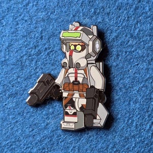 Puede incluir: Un pin con un personaje estilizado con casco y armadura blancos y grises, sosteniendo dos blásters negros. El personaje tiene una raya roja en el centro y una visera verde. El pin está sobre un fondo azul.