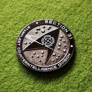 Puede incluir: Una moneda plateada de la División de Contrainteligencia de la Sección 31 de Starfleet. La moneda presenta un diseño de flecha negra con un símbolo de diana en el centro, rodeado por el texto "STARFLEET COUNTERINTELLIGENCE DIVISION" y "SECTION 31".
