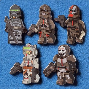 Peut inclure: Cinq épingles en émail détaillées représentant des personnages d'une franchise de science-fiction. Chaque épingle représente un soldat avec un casque et une armure uniques, tenant une arme ou un accessoire. La palette de couleurs comprend des nuances de gris, de noir, de blanc et de rouge.