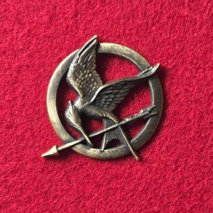 May include: A bronze-colored pin featuring a stylized bird with outstretched wings within a circular frame. The bird is positioned above two crossed arrows. The pin is set against a red background.