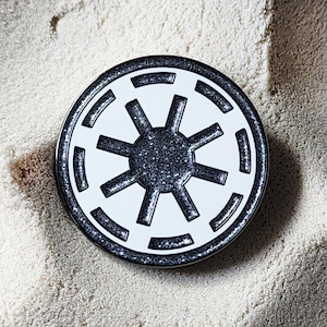 May include: A round, white and black pin featuring a stylized sunburst design. The pin has a black glittery center and a black border. The design is set against a sandy background.