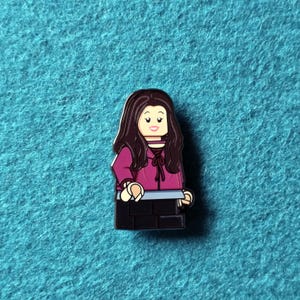 Puede incluir: Un pin estilo Lego con un personaje de dibujos animados con pelo largo castaño, que lleva una chaqueta magenta y pantalones negros. El personaje sostiene un objeto gris. El pin está sobre un fondo turquesa.