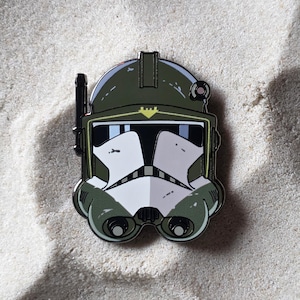May include: A detailed enamel pin featuring a stylized helmet design in shades of green, white, and black. The pin has a glossy finish and is set against a sandy background. The helmet has a distinctive visor and antenna.