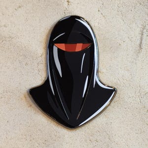 Può includere: Una spilla in smalto nero lucido a forma di casco stilizzato con una parte superiore arrotondata e una base appuntita. Il design presenta un'unica apertura orizzontale per la visiera che rivela una sezione interna di colore arancione e rosso.