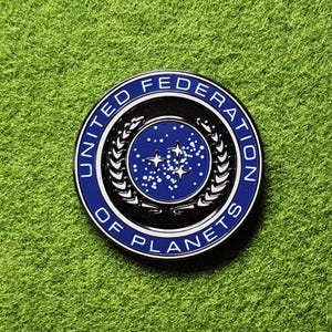 Op de afbeelding: Een ronde, blauw, zwart en witte pin met de tekst "UNITED FEDERATION OF PLANETS" rondom de rand. Het midden heeft een blauw veld met sterren en een gestileerd embleem.