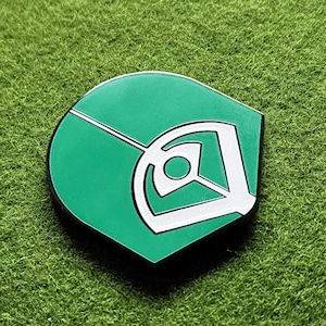 May include: A green and white enamel pin in the shape of a baseball field. The pin features a green background with white lines and a white design in the centre. The pin is set against a green background.