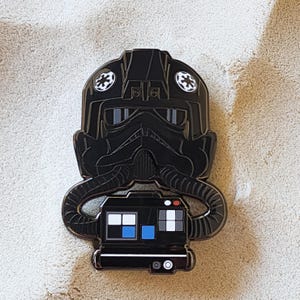 Könnte beinhalten: Schwarzer Emaille-Pin mit einem TIE-Jäger-Pilotenhelm und einem Bedienfeld. Der Pin hat weiße und blaue Akzente sowie zwei weiße runde Embleme. Ein Sammlerstück.