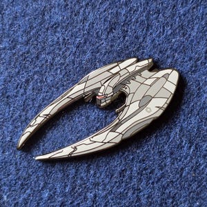 May include: A metal enamel pin shaped like a crescent-winged spacecraft. The design features off-white and light gray panels with black outlines, a central cockpit area with a small red horizontal line, and a small hexagonal emblem on the wing.