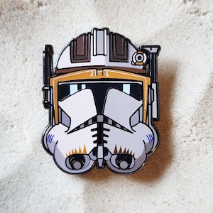 May include: A metal enamel pin shaped like a white, yellow, and brown clone trooper helmet. The helmet features a black visor, a central antenna on the left side, and distinct geometric markings in yellow and brown across the forehead and chin.