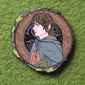 May include: A round enamel pin featuring a side profile of a character from The Lord of the Rings. The pin has a brown border with elvish script, a blue and brown background, and green accents. The character is wearing a gray cloak.