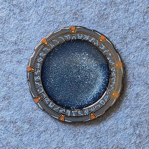 May include: A circular pin featuring a detailed design with a gray outer ring, orange accents, and a blue center with glitter. The pin has intricate symbols around the edge, resembling a portal or stargate.