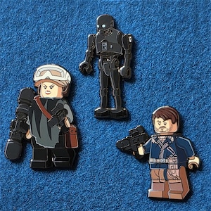 May include: Three enamel pins featuring characters from a science fiction film. The pins depict a female character with a white helmet, a black robot, and a male character in a blue jacket, all in a block figure style.