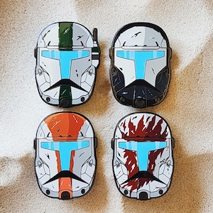 以下が含まれることがあります： ヘルメットデザインのAirPodケース4つ。各ケースは、緑とグレー、黒とグレー、オレンジとグレー、赤と白の異なる配色が特徴です。すべてのケースには青いバイザーのデザインがあります。