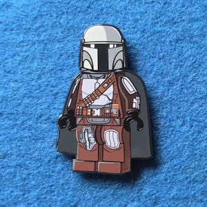 May include: A Mandalorian character pin in a Lego-style design. The pin features a helmet, armor, and cape in shades of gray, brown, and black. The pin is set against a blue background.