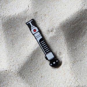 Puede incluir: Un pin en forma de sable de luz negro, blanco y gris. El pin tiene un botón rojo y una serie de líneas verticales. El pin está sobre una superficie arenosa.