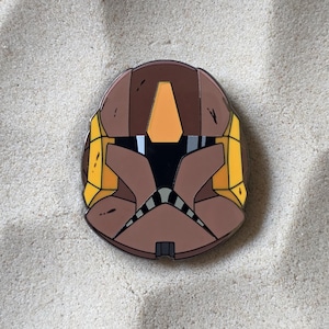 May include: A brown, yellow, and black enamel pin depicting a stylized helmet. The pin features a brown base with yellow and black accents, and a black visor. The design is symmetrical and detailed, with clean lines and a glossy finish.
