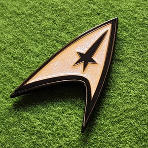 May include: A gold and black Starfleet command badge pin, shaped like a stylized arrowhead, with a black star in the center. The pin is set against a green, textured background.