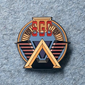 Puede incluir: Un pin redondo de esmalte con un diseño geométrico y las letras "SGC" en rojo. El pin presenta una combinación de colores dorados, azules y negros con formas triangulares y de alas. El pin está sobre una superficie de fieltro azul.