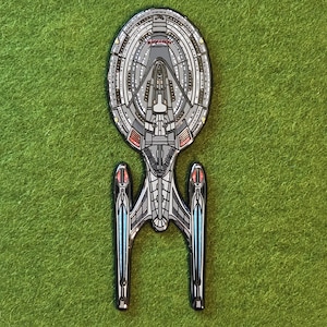 Puede incluir: Un pin de esmalte metálico con la forma de la nave espacial USS Enterprise-E de Star Trek. El diseño presenta un casco gris con detalles complejos de paneles negros, acentos rojos en las barquillas y reflejos azules. El texto U.S.S. ENTERPRISE es visible.