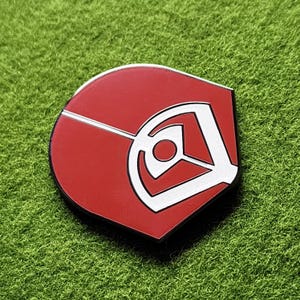 May include: A red and white enamel pin with a distinctive geometric design. The pin has a red background with a white outline and a white geometric shape in the centre. The pin is set against a green background.
