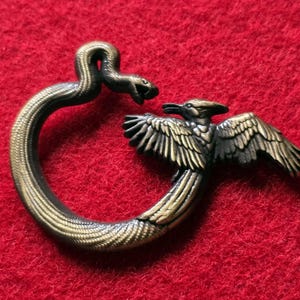 May include: A bronze metal pendant depicting a snake biting a bird. The snake is curved, and the bird has outstretched wings. The pendant is set against a red background.