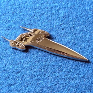Puede incluir: Un broche de metal plateado pulido con forma de nave espacial puntiaguda y elegante. El diseño presenta una nariz larga y cónica, un cuerpo central con detalles geométricos grabados y dos protuberancias en forma de ala orientadas hacia atrás.