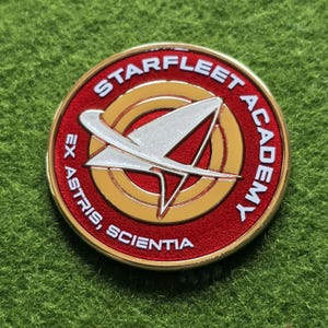Puede incluir: Un pin redondo rojo y dorado de la Academia Starfleet. El pin presenta las palabras "STARFLEET ACADEMY" en blanco y "EX ASTRIS, SCIENTIA" en blanco. Un emblema plateado y blanco está en el centro.