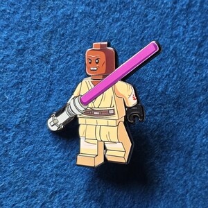 Peut inclure: Épingle représentant une figurine avec une tête marron et une tenue beige, tenant un sabre laser violet. La figurine a un emblème rouge sur l'épaule et un bras argenté et noir. Fond bleu texturé.