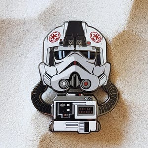 May include: Enamel pin depicting a white and black AT-AT pilot helmet with red details and a control panel. The pin has a detailed, stylised design with a silver outline. The background is a neutral colour.