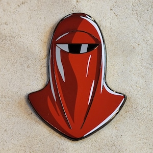 May include: A red enamel pin shaped like a stylized helmet with a smooth, glossy finish. The design features a pointed, rounded top, a wide central visor with a black horizontal slit, and white highlights that create a sense of depth and dimension.