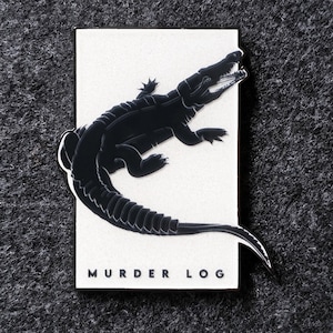 May include: A rectangular enamel pin featuring a white background with a black silhouette of an alligator. The alligator is depicted with its mouth open, and the text "MURDER LOG" is printed in black capital letters at the bottom of the pin.
