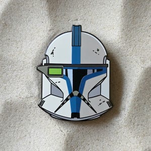 Op de afbeelding: Een wit en blauw emaille speldje in de vorm van een Clone Trooper helm. De pin heeft een blauwe streep in het midden en een groen rechthoek aan de zijkant. De helm staat tegen een zanderige achtergrond.
