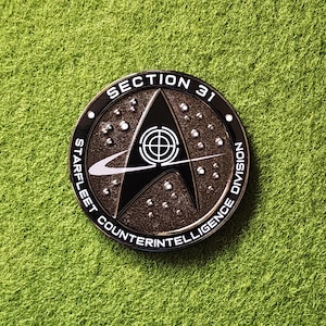 May include: A round, metallic Section 31 Starfleet Counterintelligence Division coin. The coin features a black and white emblem with the text "SECTION 31" and "STARFLEET COUNTERINTELLIGENCE DIVISION" around the edge. The coin is set against a green background.