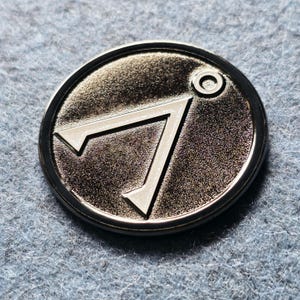 Könnte beinhalten: Eine silberfarbene, kreisförmige Metallmünze mit einem stilisierten Design. Das Design zeigt eine große, eckige Form und einen kleinen Kreis. Die Münze hat eine glänzende, reflektierende Oberfläche und einen dunklen Rand. Es ist wahrscheinlich ein Sammler- oder Dekorationsartikel.