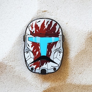 May include: An enamel pin depicting a helmet design with a white base, red and grey patterns, and a light blue visor. The pin has a black border and is set against a sandy background. The design is highly detailed.