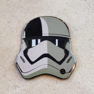 May include: A metal enamel pin shaped like a stormtrooper helmet. The design is split vertically, featuring a black and white color scheme on the left side and a muted sage green and dark gray color scheme on the right side, with black outlines.
