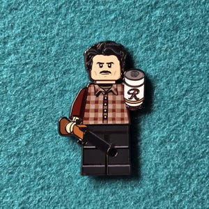 Puede incluir: Un pin estilo Lego con un personaje con una camisa a cuadros marrón y beige, pantalones negros y un bigote oscuro. El personaje sostiene una lata con una etiqueta blanca y una gran "R", y un objeto marrón que se asemeja a una escopeta.